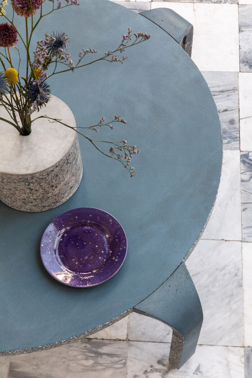 Round blue table with a granite vase holding flowers and a purple plate on a checkered gray and white marble floor.