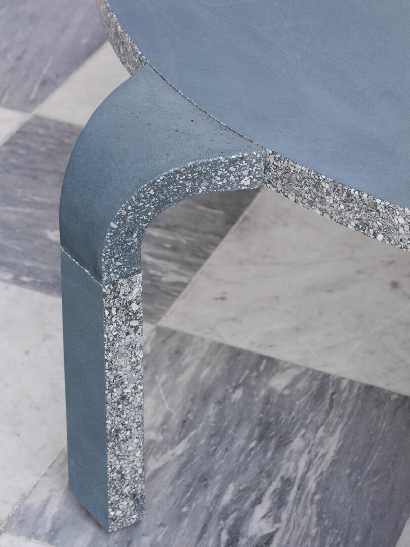 Close-up of a smooth, round blue table with terrazzo detailing on its edge and leg, placed on a checkered marble floor.