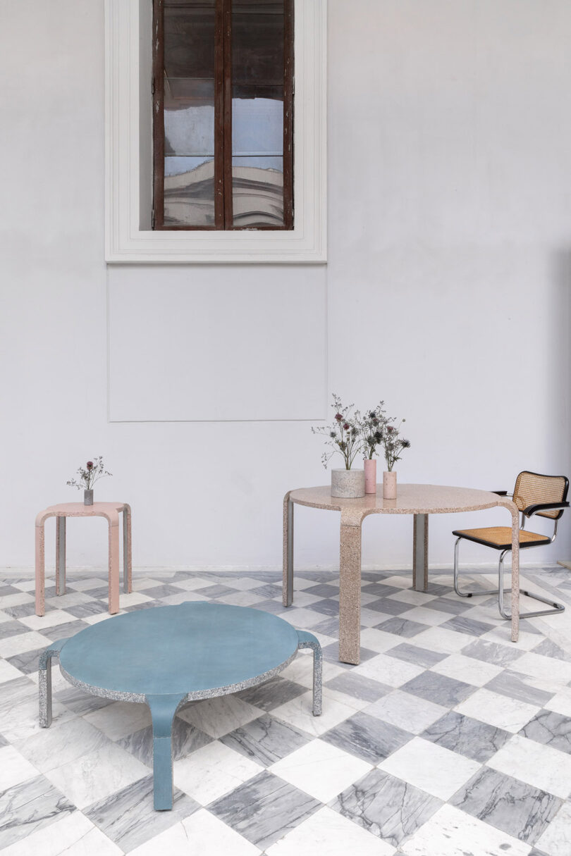 Minimalist room with three modern tables and a chair on a checkered floor. Tables hold vases with flowers. A high window is in the background.