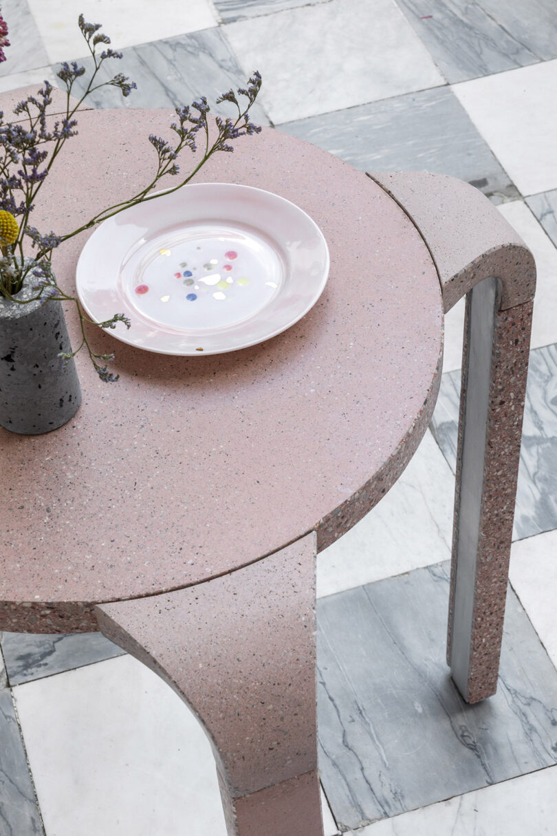 Round pink terrazzo table with metal legs, a small vase of flowers, and a plate with confetti. The floor is checkered with white and gray tiles.