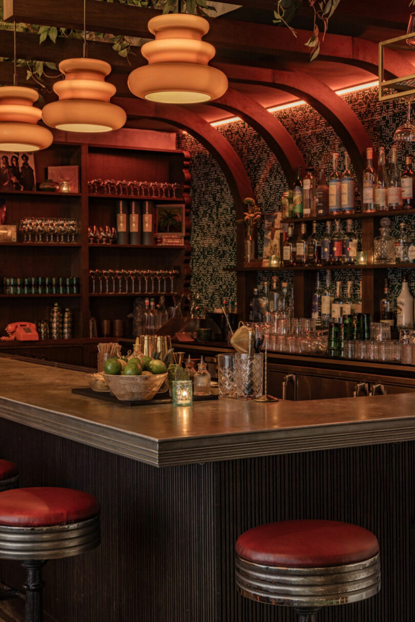 Warmly lit bar interior with shelves of glasses and bottles. Three pendant lights hang above the counter, which has a candle and a fruit bowl. Red-cushioned stools line the bar.