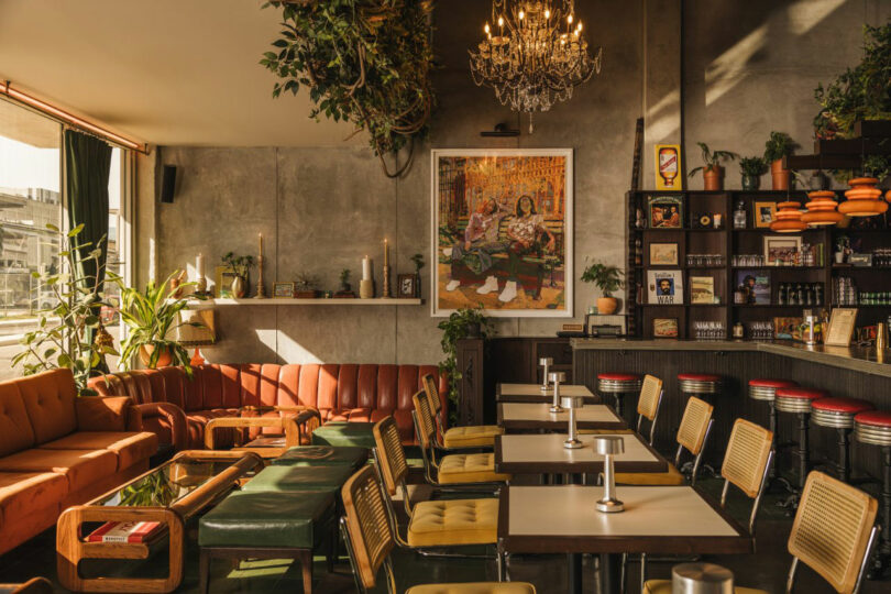 A cozy café interior with a mix of orange, brown, and green seating, plants, a chandelier, and wall art. Tables and chairs are arranged neatly next to a shelf filled with books and decor.