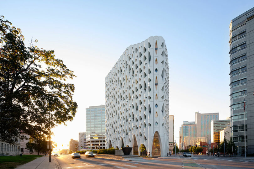 A modern high-rise building with a unique, geometric facade towers like a tree against the cityscape and a clear blue sky.