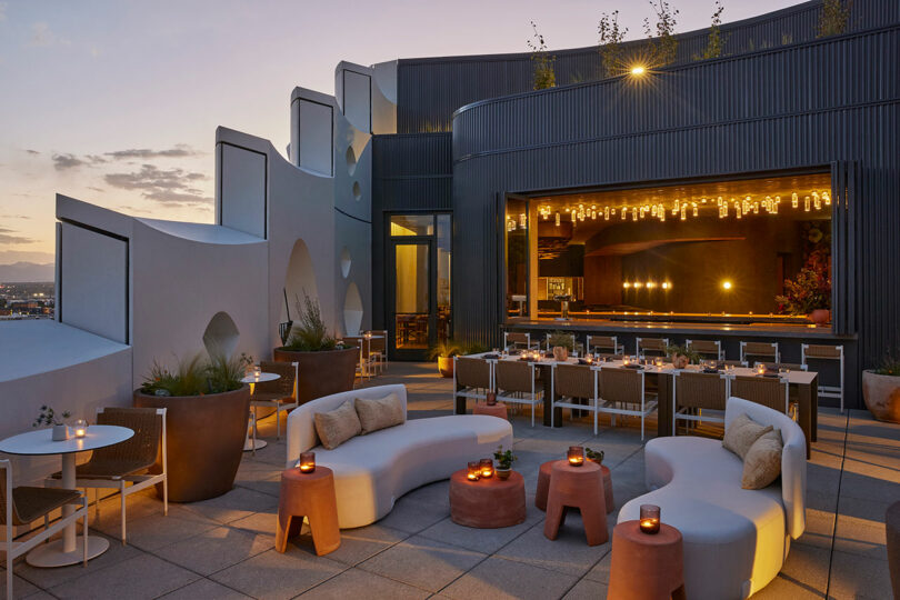 Experience the Populus rooftop bar at sunset, where modern curved seating and tables invite relaxation. Enjoy the well-lit bar area as you take in the breathtaking cityscape views in the background.