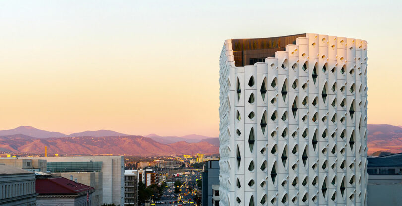 A white Populus-inspired building with a patterned facade stands tall against a cityscape, framed by mountains in the background during sunset.