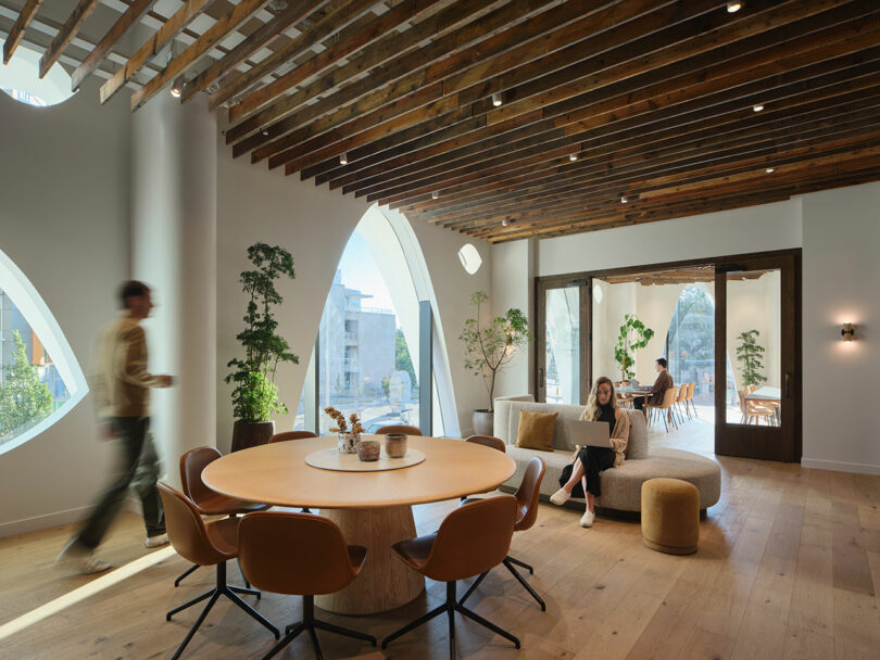 Modern office space with Populus-themed wooden decor, round table, chairs, and sofas. A person walks past while another works on a laptop. Large arched windows provide natural light and exterior views.