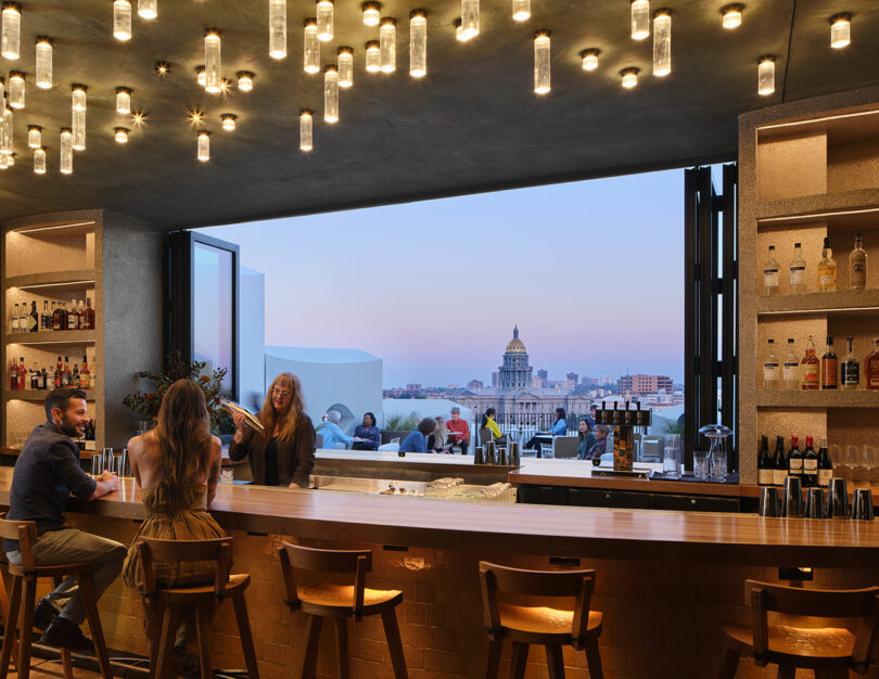 Rooftop bar with a cityscape view. Several patrons are seated at a wooden counter under hanging lights, enjoying Populus cocktails. A large window frames the distant city buildings under a twilight sky.