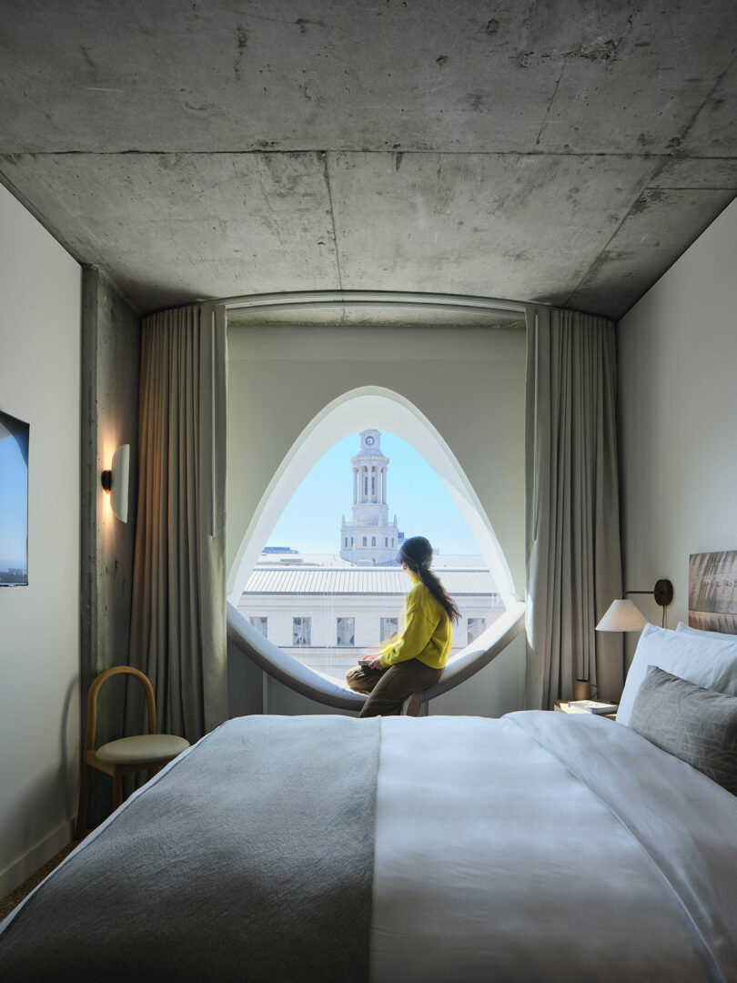 A person in a vibrant yellow shirt sits on the window ledge of a modern Populus hotel room, gazing thoughtfully at a distant clock tower.