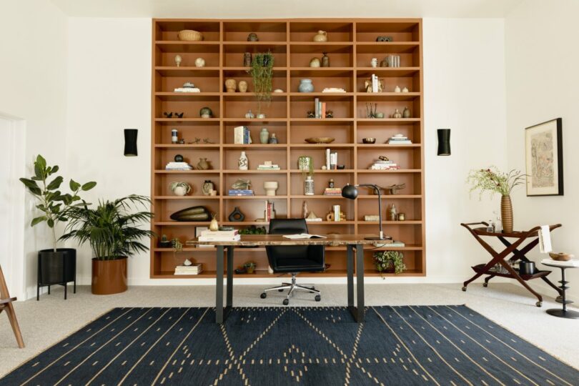 A modern office features a large wall-mounted wooden bookshelf, a desk with a black chair, a blue patterned rug, and various plants and decorations.