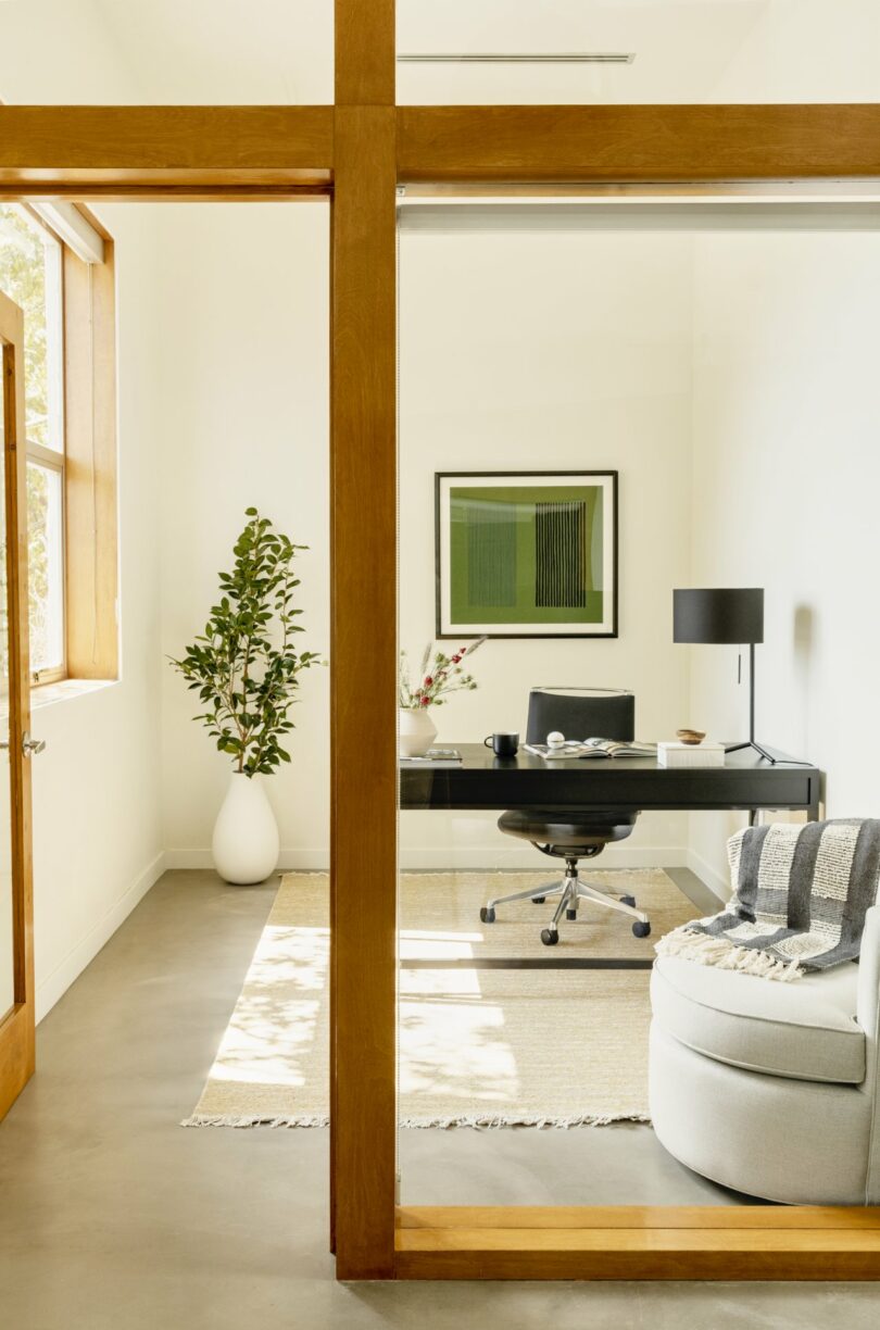 Modern office with a black desk, chair, and lamp. A potted plant is by the window. A framed abstract artwork is on the wall. There is a round chair with a striped blanket in the corner.