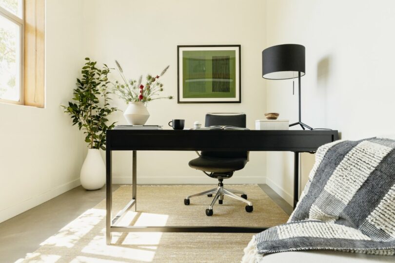 Minimalist home office with a black desk, chair, and lamp. Decorative vase with plants is on the desk. A framed artwork and potted plant are visible. There's a striped throw on a nearby chair.