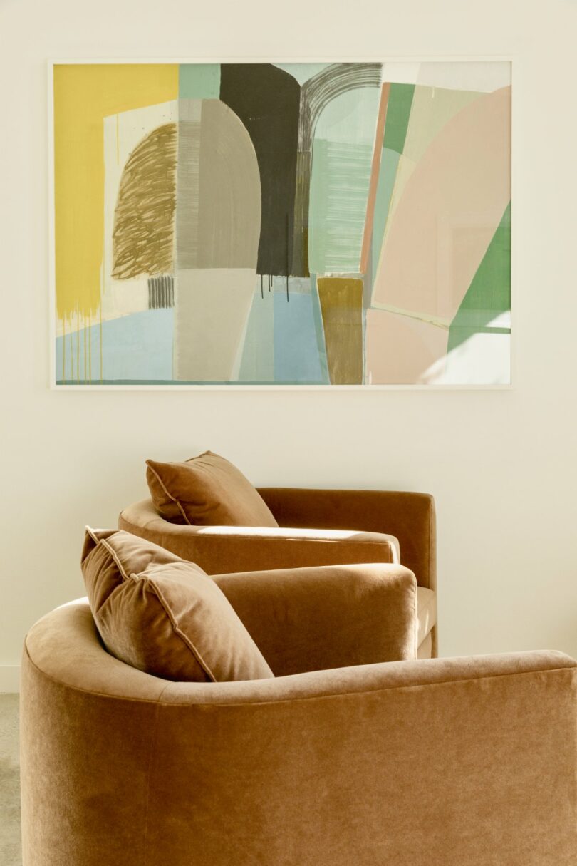 Two tan velvet chairs face an abstract painting with geometric shapes and various colors, including green, beige, and yellow, mounted on a light-colored wall.