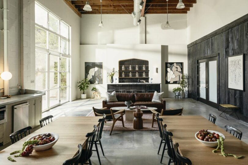 A modern living and dining area with high ceilings, a wooden dining table, chairs, a brown sofa, and large windows. Artworks and plants decorate the space.