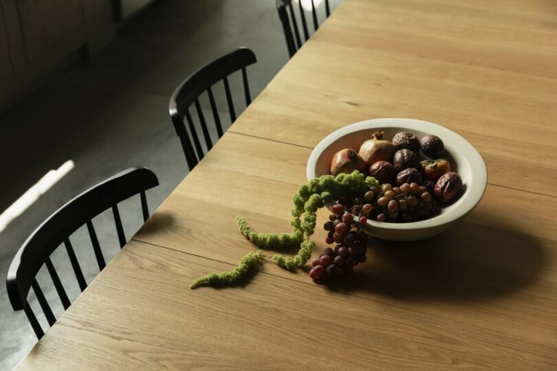 A wooden table with black chairs, holding a bowl of assorted fruits, including grapes, dates, and a pomegranate, with some grapes spilling onto the table.