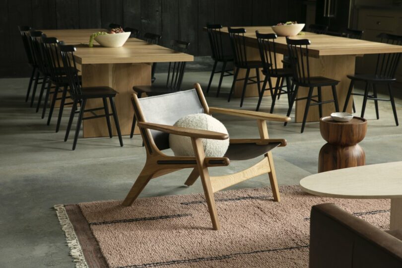 A modern, light wood chair with a gray cushion and a white pillow sits in a stylish room. Nearby are wooden tables with black chairs and small tables on a textured rug.