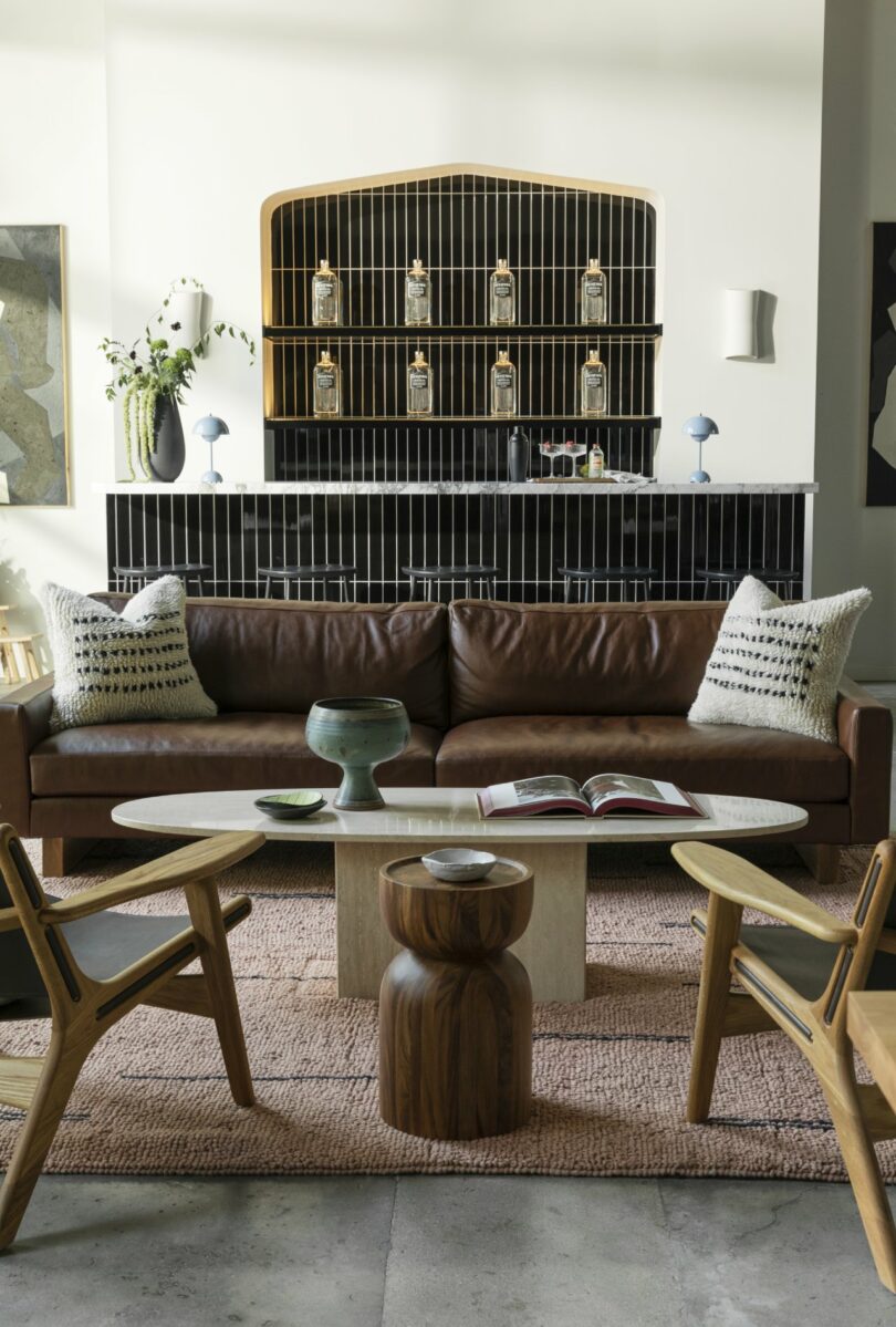 Cozy living room with a brown leather sofa, decorative pillows, two wooden chairs, a round coffee table, and a backlit shelf featuring decorative items. Neutral tones and natural textures dominate.