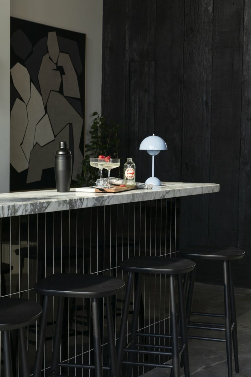 Modern bar area with a marble countertop, four black stools, a cocktail shaker, two drinks with berries, a lamp, a plant, and an abstract wall art piece.