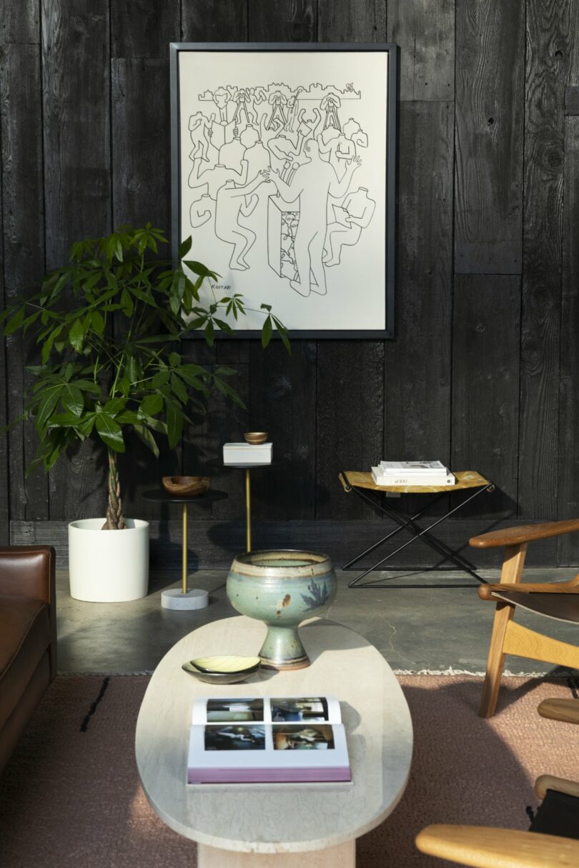 A cozy living room with a modern design, featuring a leather chair, a small table with books, a potted plant, a framed abstract artwork on a dark wood wall, and additional seating.