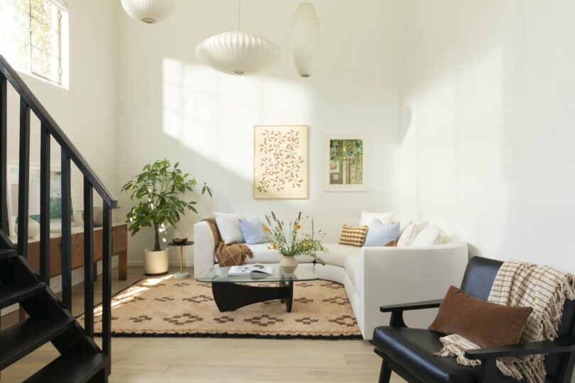 A living room with a white sectional sofa, patterned rug, and a coffee table. There are plants, wall art, and three pendant lights. Black stairs and wooden floor are also visible.