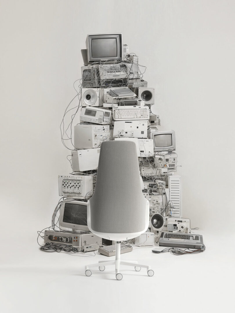 A tall stack of vintage electronic equipment and computers with tangled wires evokes a nostalgic charm, as if inviting someone to take five in the modern swivel chair, embracing both past and present technology.