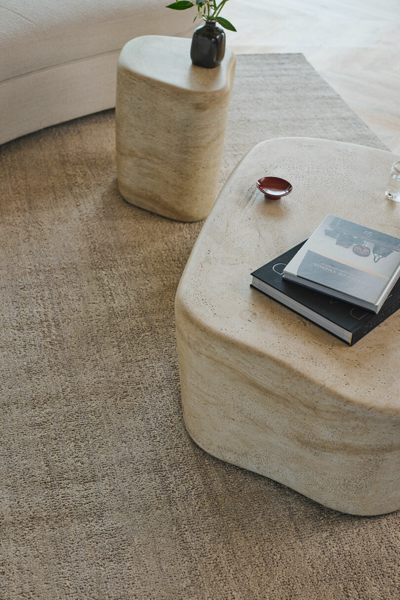 Two beige stone tables on a light brown rug with a vase, a red dish, and books placed on top