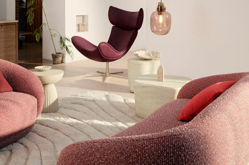 Modern living room with a burgundy lounge chair, textured sofa, and round tables on a patterned rug. Natural light and minimalist decor enhance the space