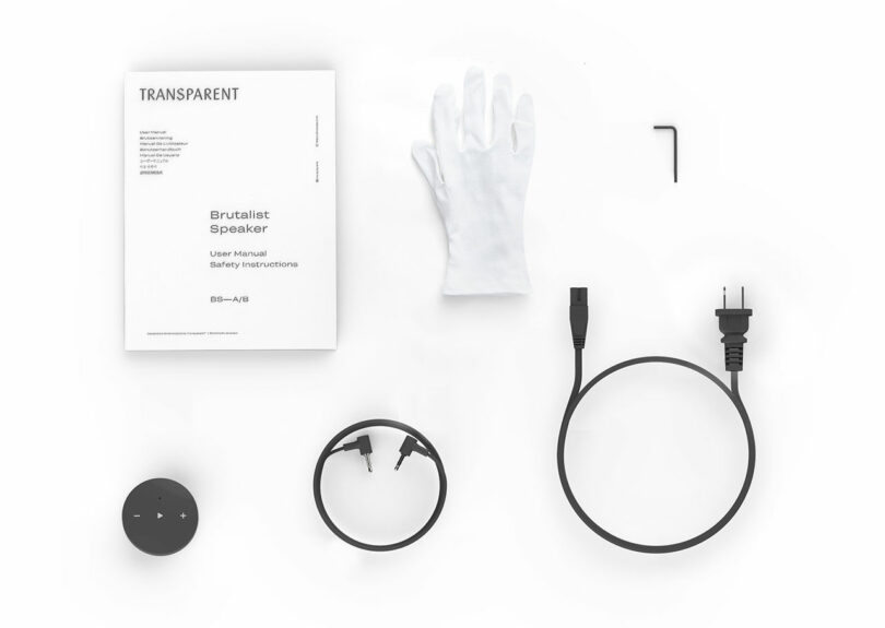 Brutalist audio speaker assembly kit featuring a manual, white gloves, Allen key, power cable, audio cables, and a control knob on a stark white background.