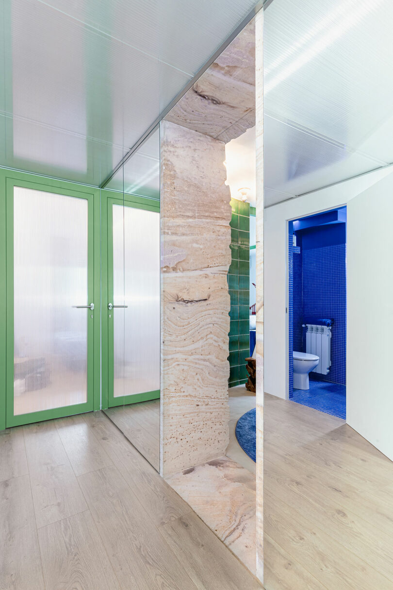 Un couloir moderne avec un parquet en bois clair, des portes vitrées à cadre vert et une cloison à texture de pierre naturelle. Un aperçu d’une salle de bains au carrelage bleu est visible à travers une porte ouverte.