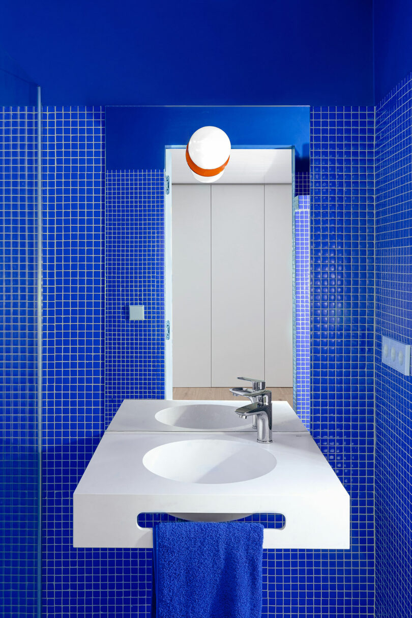 Salle de bains avec murs carrelés bleus, comptoir blanc à double vasque et serviette bleue. Un plafonnier rond est visible près de l'entrée.