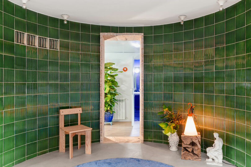 Chambre avec murs carrelés verts, chaise en bois, plantes, lampadaire et statue. Une porte centrale mène à un espace avec plus de plantes et un mur bleu.