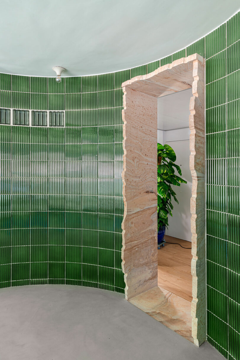 A room with glossy green tiles and a rough stone doorway leading to another space with a plant in the background.