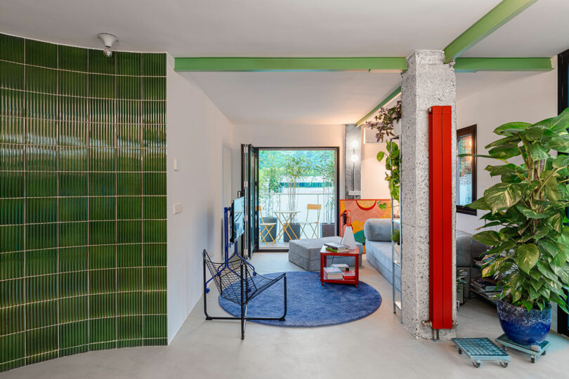 Salon moderne avec mur d'accent carrelé vert, tapis circulaire bleu et mélange de meubles contemporains. De grandes plantes en pot et un petit balcon sont visibles en arrière-plan.