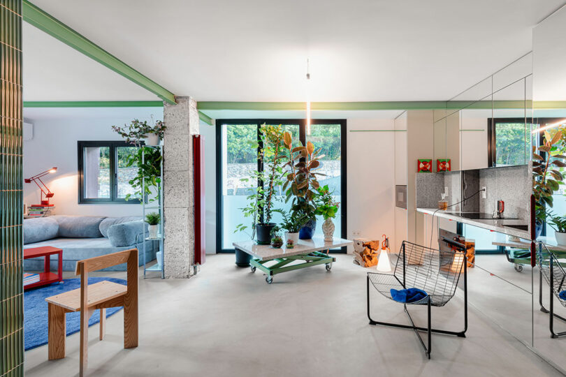 Salon moderne avec un mélange de meubles dont un canapé bleu, une chaise en bois et une chaise en fil de fer. Les plantes d'intérieur décorent l'espace et les grandes fenêtres apportent de la lumière naturelle.