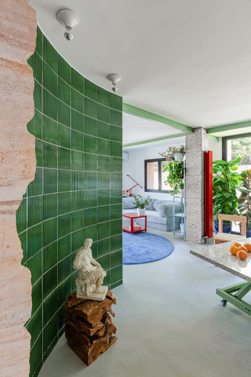 Scène d'intérieur avec un mur carrelé vert, une statue sur un socle en bois, un tapis rond bleu et un mobilier moderne. De grandes plantes sont près des fenêtres et un bol d'oranges est sur la table.