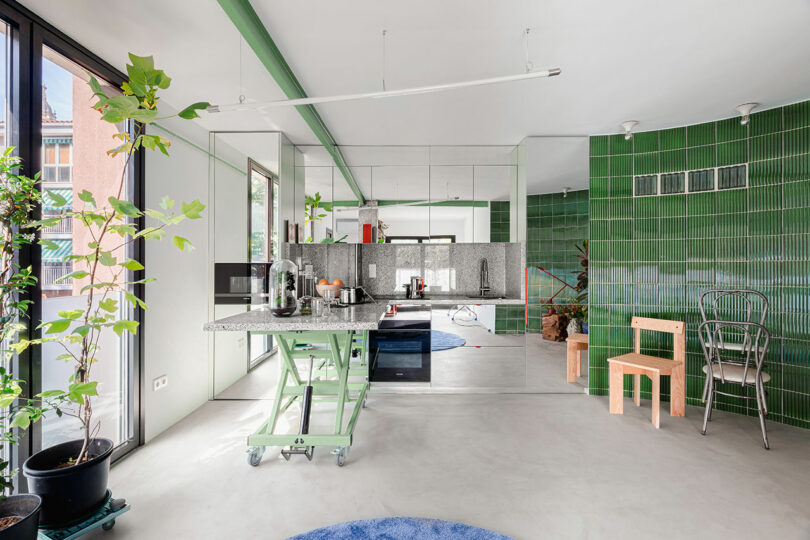 Modern kitchen with green tiles, stainless steel appliances, and potted plants. A unique adjustable-height table is central, with wooden and metal chairs nearby. Large windows provide natural light.