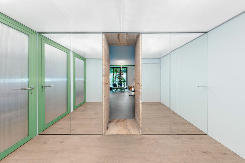 Un couloir moderne avec des portes vitrées aux cadres verts sur les côtés, du parquet en bois clair et une ouverture centrale menant à une pièce avec des plantes.