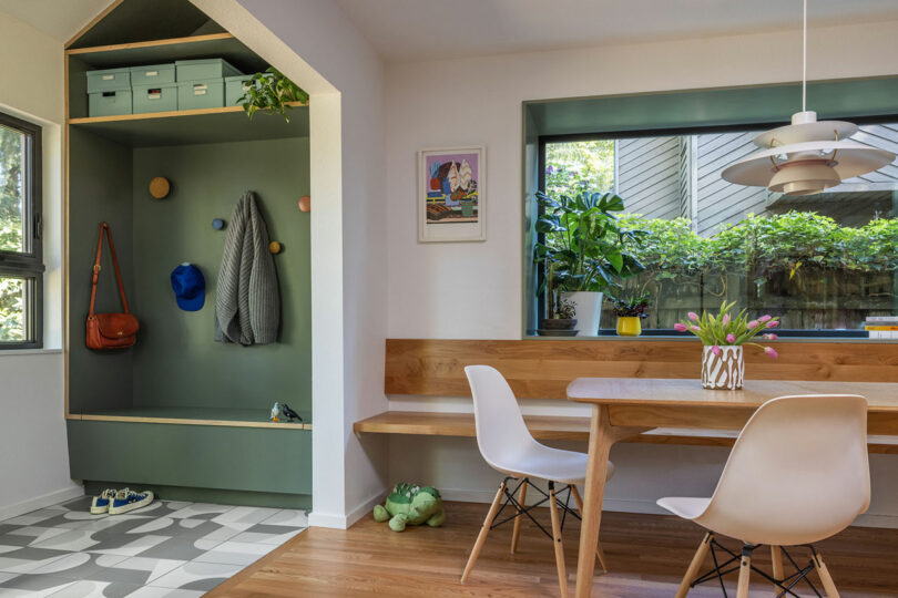 Un acogedor comedor con una mesa de madera, sillas blancas y un asiento de ventana con plantas. Un cubero verde incorporado con ganchos, estantes y un banco está cerca. La luz natural ilumina el espacio.