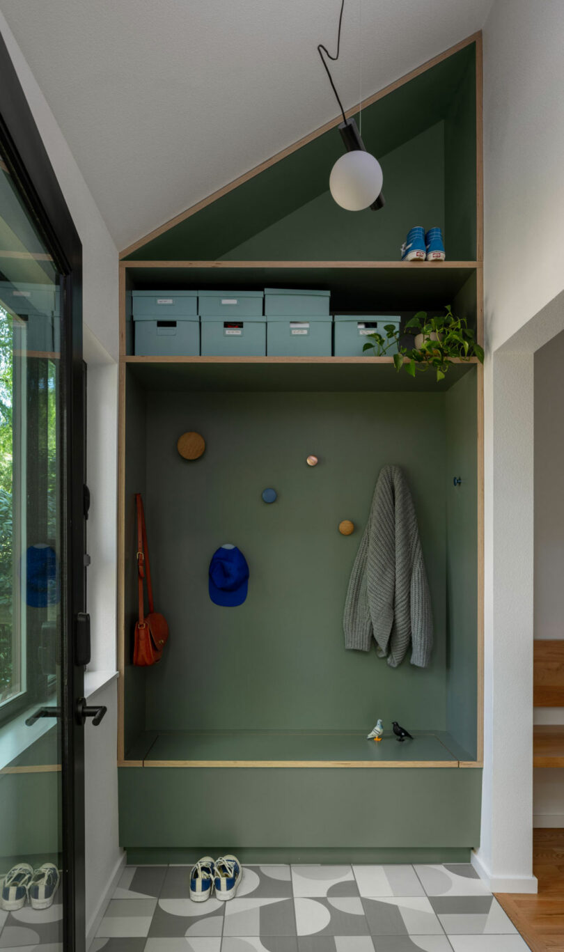 A green entryway nook with shelves, hat and coat hooks, assorted boxes, shoes, a hanging gray sweater, a blue cap, wall art, and a potted plant. Modern pendant light above.