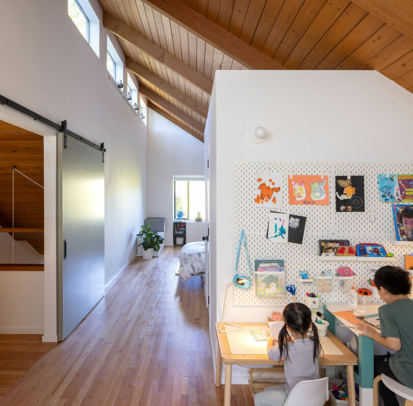 Dos niños se sientan en un dibujo de escritorio, rodeado de coloridas obras de arte en una habitación brillante con pisos de madera y un techo alto y en ángulo.