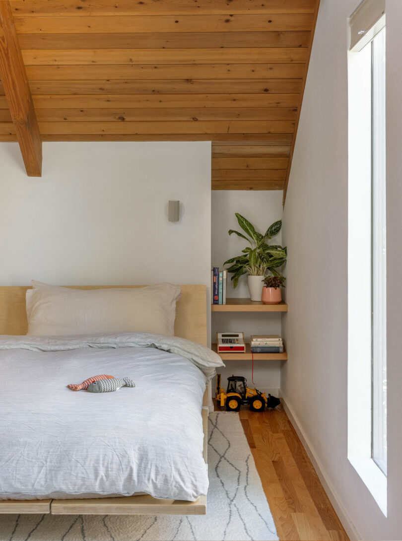 Acogedor dormitorio con techo de madera, cama con ropa de cama gris claro, juguete relleno, estantes laterales con plantas y libros, pequeños juguetes de construcción en el piso y una ventana.