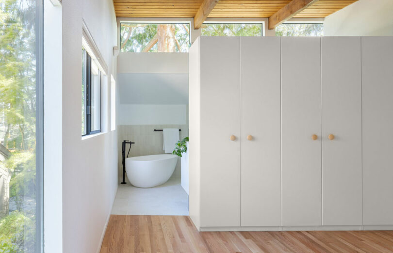 Baño moderno con una bañera blanca independiente, grifo negro y ventanas grandes. Los pisos de madera clara y los gabinetes minimalistas de gris claro son visibles. Los árboles se ven a través de las ventanas.