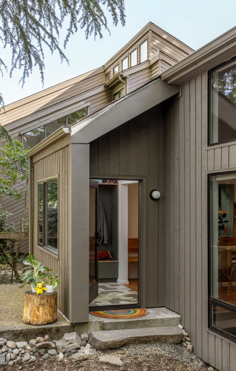 Exterior moderno de la casa con un diseño geométrico, con paneles de madera, un pequeño porche con una lámpara redonda y una alfombra colorida dentro de la entrada. La vegetación rodea la casa.