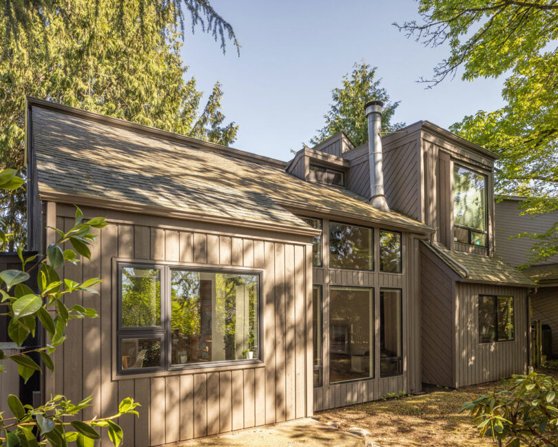 Moderna casa de dos pisos con grandes ventanas, revestimiento de madera y rodeada de árboles debajo de un cielo claro.