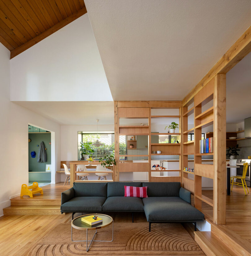 Modern living room with a green sectional sofa, wooden partition, and circular coffee table. Open plan includes dining area with white chairs, and a kitchen in the background. Bright and airy space.