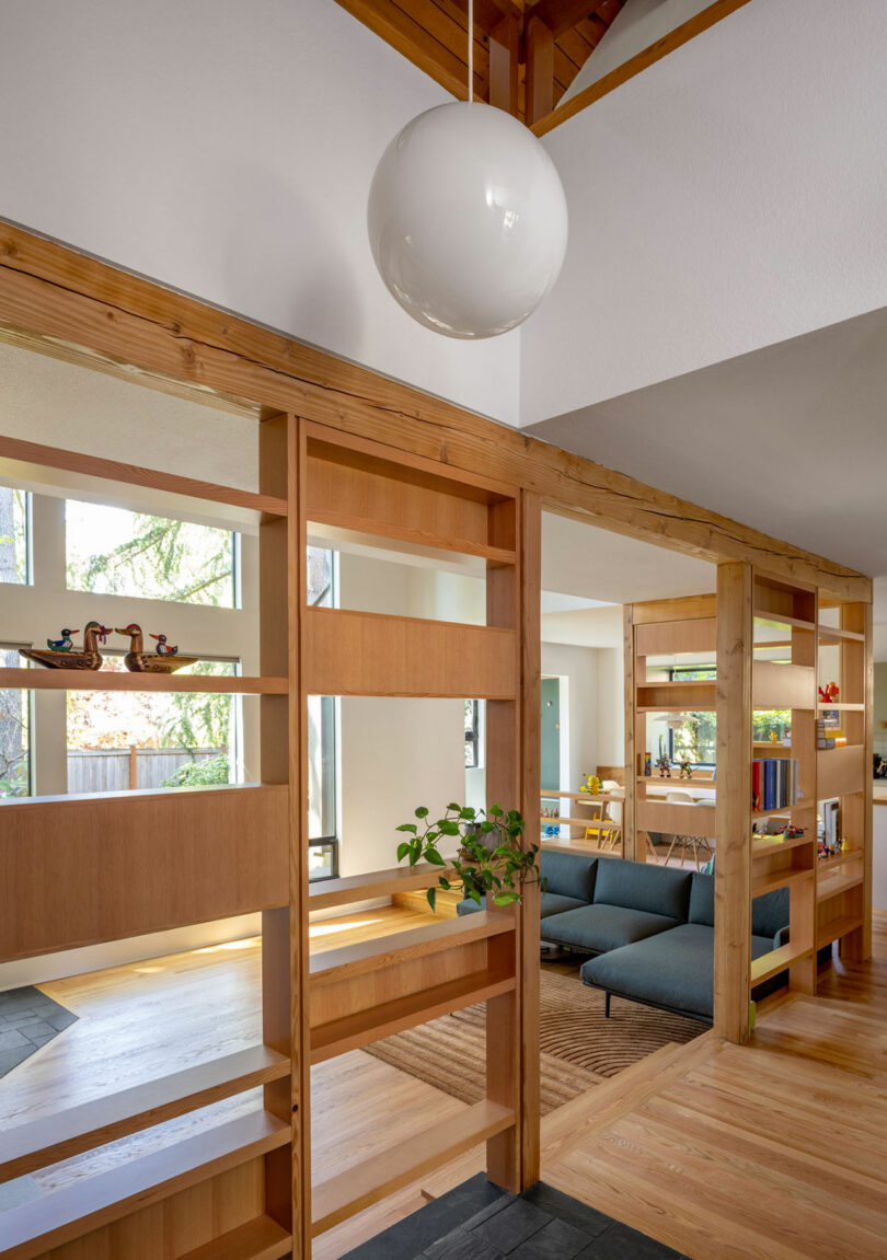 A modern living room with wooden shelves, a globe pendant light, and a potted plant. Blue sofas are visible in the background against large windows.