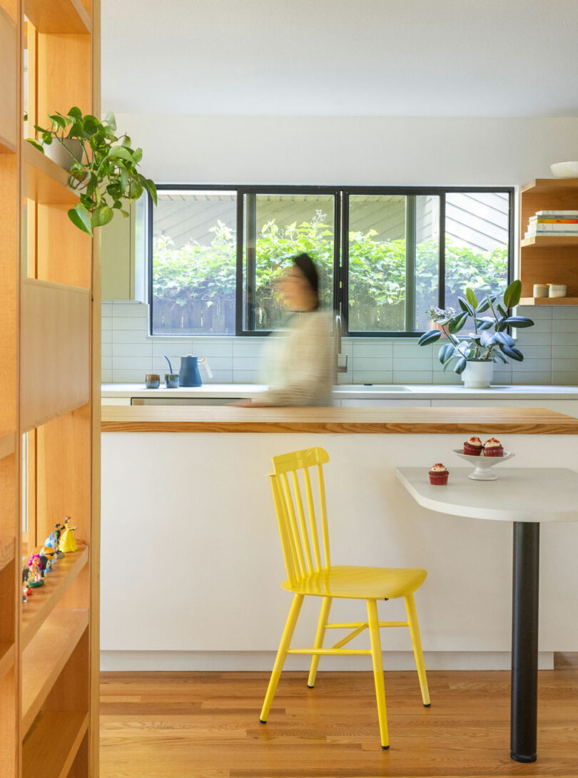 Cocina moderna con una silla amarilla en una pequeña mesa, cupcakes en la mesa, estante abierto con decoración y una persona borrosa cerca del fregadero. Ventanas grandes con vegetación en el fondo.