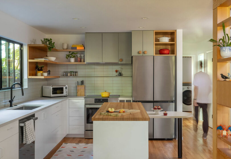 Cocina moderna con gabinetes grises y blancos, electrodomésticos de acero inoxidable y una isla de madera. Una persona camina en el fondo cerca de una zona de lavandería.