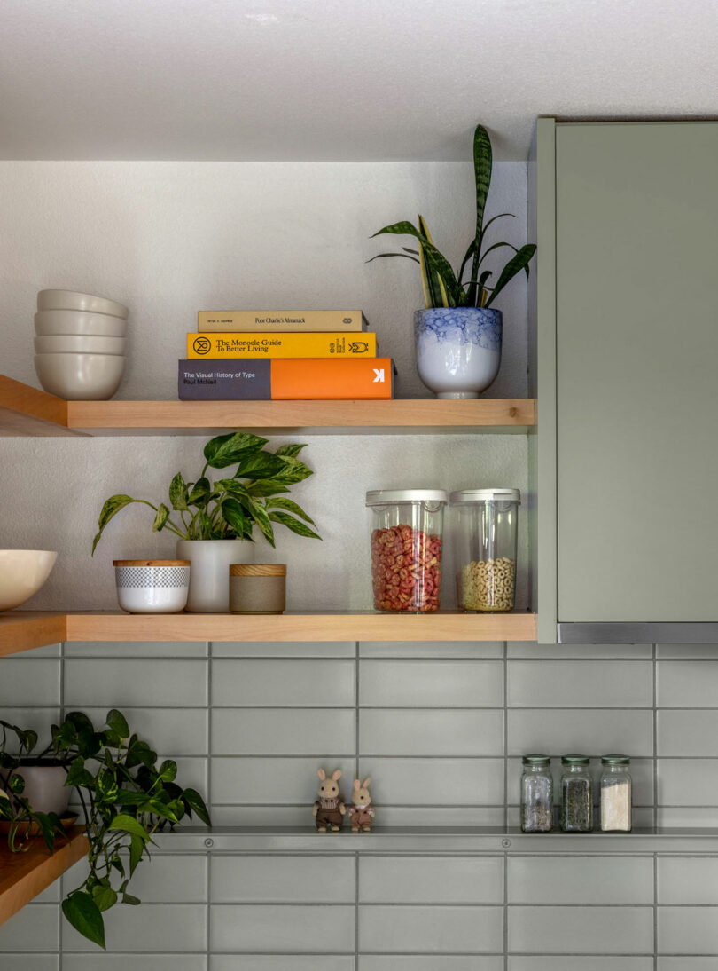 Un área de cocina con gabinetes verdes y estantes de madera que sostienen plantas, libros, tazones y frascos de frijoles y granos. Dos figuras de conejo se sientan en el mostrador.