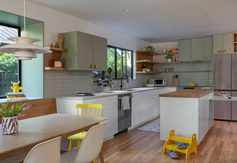 Cocina moderna con gabinetes blancos, encimeras de madera y electrodomésticos de acero inoxidable. Un comedor con sillas amarillas y blancas está cerca, y hay plantas y artículos decorativos en los estantes.