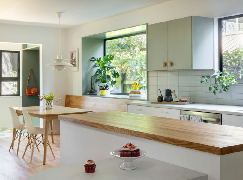 Cocina moderna con gabinetes de color verde claro, encimeras de madera, ventanas grandes y plantas de interior. Una mesa con sillas y un mostrador con dos cupcakes están en primer plano.